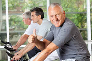 Photo of three men on exercise bikes with the closest one smiling and giving a thumbs up