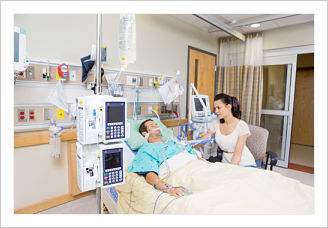 A photo of a woman sitting beside a man with medical equipment hooked up to him laying in a hospital bed