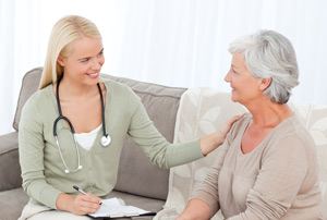 Photo of a young woman wearing a stethoscope sitting on a couch beside an elderly woman and comforting her.
